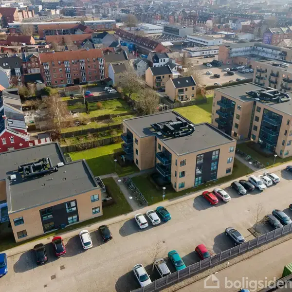 Flere moderne bygninger står stille omgivet af grønne plæner og parkeringspladser med biler. I baggrunden ses en tætbygd by med rødstenshuse og gader.
