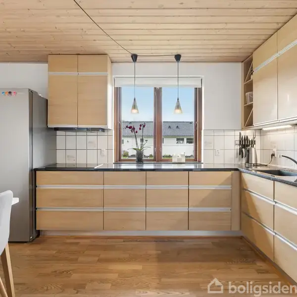 Køkken med lyse træskabe og indbygget ovn, bordplade ved vask, hængende lamper. Vindue med udsigt til huse. Stål køleskab og hvid væg. Trægulv og loft.
