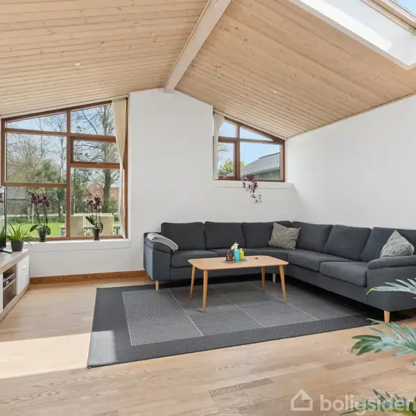 En grå sofa står i en lys stue med trægulv og skråt loft. Et sofabord er foran, og store vinduer lader naturligt lys strømme ind. En fladskærm står på et lavt skab.