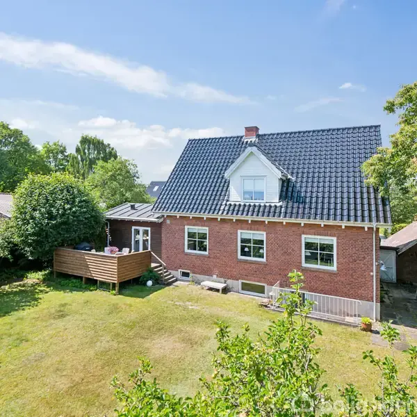 Et rødt murstenshus med sort tag står i en grøn have. En terrasse ses til venstre, omgivet af buske og græs, under en klar blå himmel.