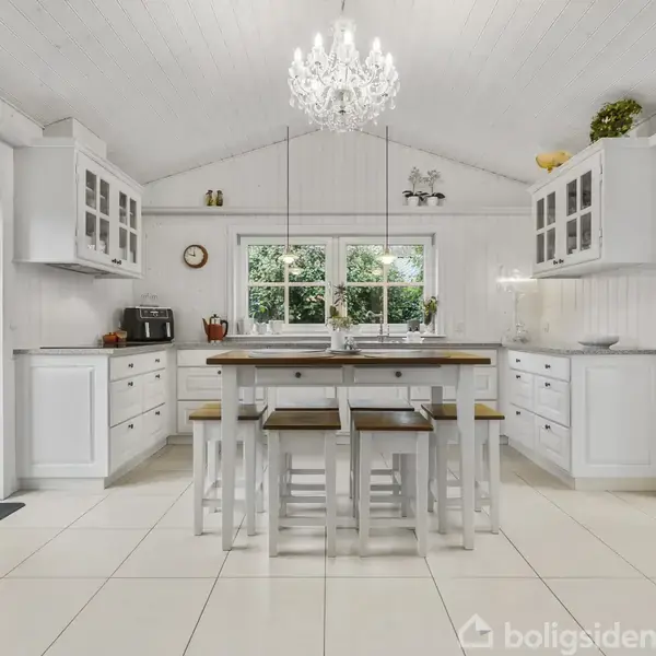 Et lyst køkken med hvidt interiør har et centralt spisebord med trætop. Omkring bordet står hvide stole. Over bordet hænger en lysekrone. Vinduer viser en grøn have.