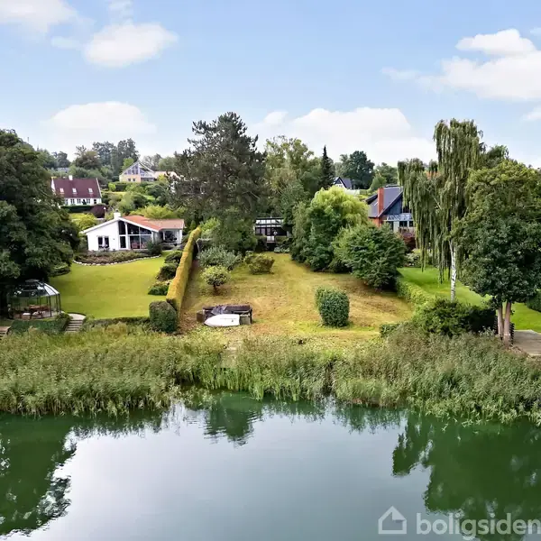 Bolig ved søen med have, omgivet af træer og buske. Hvide huse i baggrunden, små bådebroer ved vandkanten. Græsplæne strækker sig fra hus til sø.