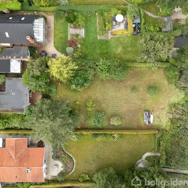 Huse set fra oven med haver, stier og træer omgivet af en sø til højre. Græsarealer og terrasser skaber en harmonisk haveindretning blandt bebyggelserne.