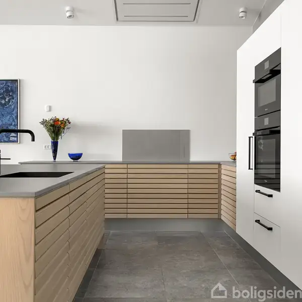 Køkken med lyse træskabsfronter og sort bordplade; en moderne ovn i væggen. En vandhane stikker op fra øen. Maleri og blomsterdekorationer skaber en elegant atmosfære.