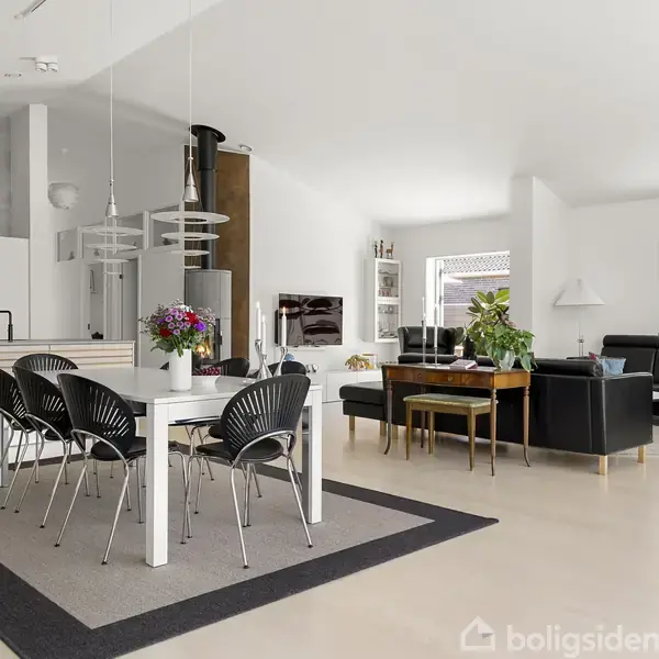 Spisebord med sorte stole står på et tæppe i et moderne, lyst køkken-alrum. I baggrunden ses en stue med mørke sofaer og store vinduer, samt planter og dekorationer.