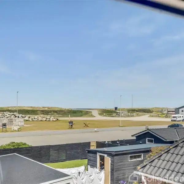 Et forstadsområde med hustage i forgrunden, en åben vej, og græsbeklædte klitter i baggrunden under en klar, blå himmel.