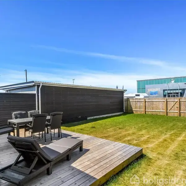 En træterrasse med liggestol og spisebordssæt står i en baghave. Græsplæne omgiver terrassen. I baggrunden ses en træhegn og en stor bygning under en blå himmel.