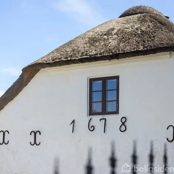 Hus med stråtag og lille vindue har teksten "1678" og dekorative detaljer på den hvide facade; omgivet af blå himmel og grøn vegetation.