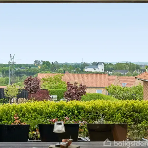 Ydersigt over en terrasse med potteplanter, der står stille. I baggrunden ses bygninger med røde tage, grønne buske og træer samt en lys, klar himmel.