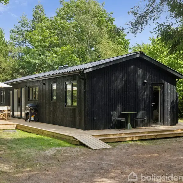 Sort træhus står stille på en træterrasse i en skovklædt omgivelser. En parasol og havemøbler er placeret på terrassen, og frodige grønne træer omkranser området.