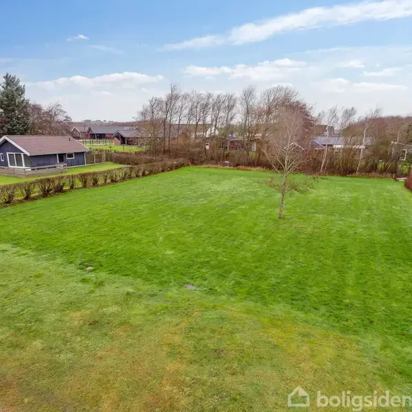 Et gråmalet hus står på en grøn græsplæne omgivet af hække. Det er en klar dag med blå himmel, og træer uden blade ses i baggrunden.