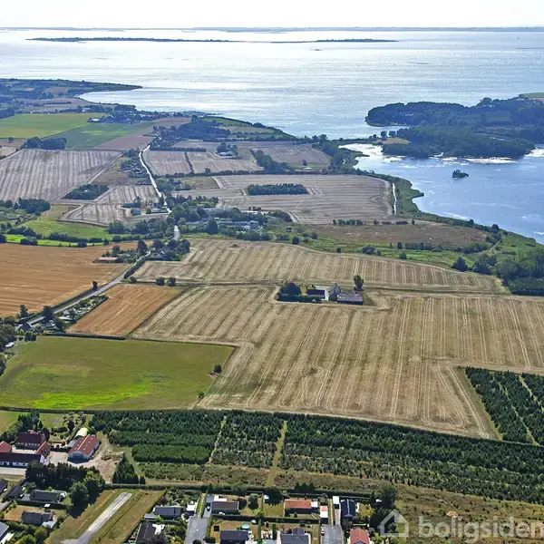 Luftfoto viser marker og små landsbyer med en vej, der strækker sig gennem landskabet, nær kysten med udsigt til havet i baggrunden. En pil peger mod et hus.