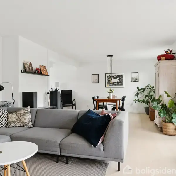 Grå sofa står i en stue ved et rundt hvidt sofabord, omgivet af planter og kunst på væggene. Spisebord og stole ses i baggrunden under en hængelampe.