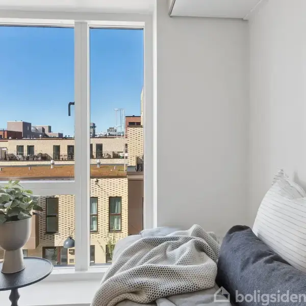 En lys stue med en stor vindue, der viser en udsigt til byens tage. En sofa med puder og et tæppe står ved siden af et lille bord med en potteplante.