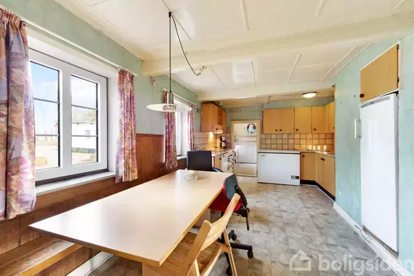 Et køkken med et langt spisebord, stole og en bænk står under et vindue til venstre. Køkkenskabe, opvaskemaskine og komfur danner en funktionel arbejdszone i baggrunden.