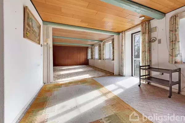 Rummeligt værelse med trægulv og træloft, oplyst af sollys gennem store vinduer. Maleri på væggen og en åben dør til en terrasse. Ingen tekst i billedet.