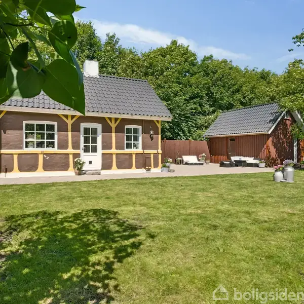Lille bindingsværkshus med gråtag, placeret i en solrig have med græsplæne. Tilstødende er et rødt skur, omgivet af tætte grønne træer og buske under en klar blå himmel.