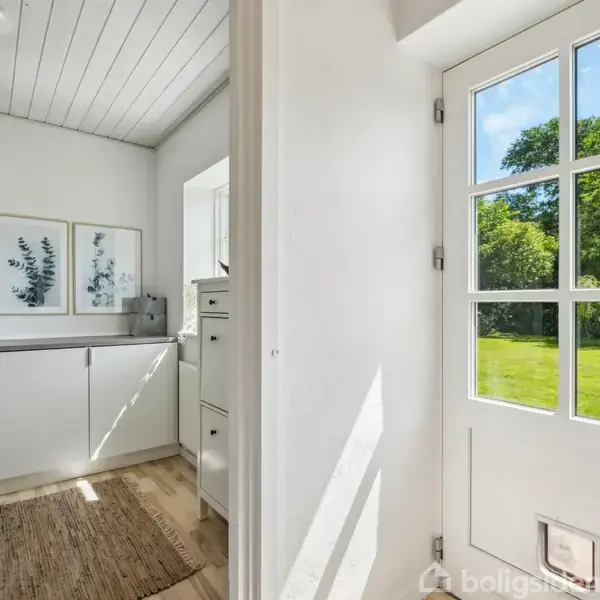 Dør åbner mod en grøn have i sollys. Indenfor er der lyse, moderne skabe, dekorative plantebilleder og en tæppebelagt trælaminatgulv.