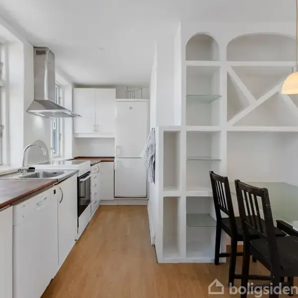 Et lyst køkken med hvide skabe og træbordplade, hvor et glasbord med sorte stole står ved siden af en hvid reol. Vinduer byder naturligt lys indenfor.