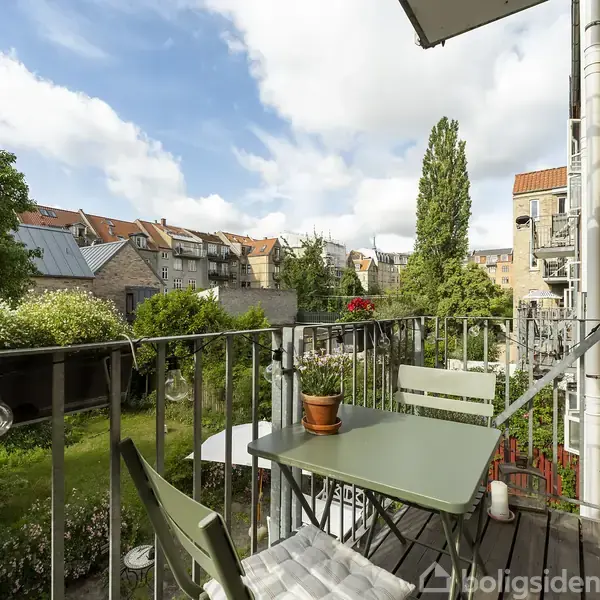 En lille altan med to stole og et bord med en plante står mod en baggrund af en grøn have og historiske bygninger under en blå himmel med skyer.
