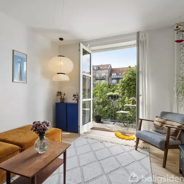 En stue med en gul sofa, sofabord med vase, lænestol og TV. Dør står åben til altan med udsigt til grønne omgivelser. Moderne og lyst indrettet.
