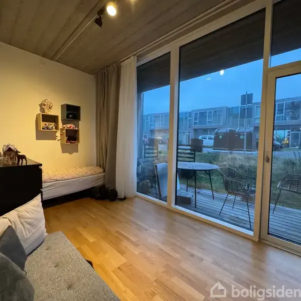 Værelset har en enkeltseng, sofa, og mørk kommode. Udenfor ses en terrasse med bord og stole. Store vinduer viser moderne boliger udenfor.