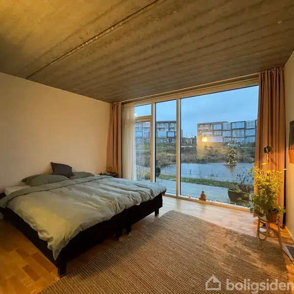 Seng står i en moderne soveværelseindretning med store vinduer, der udsigt til en baggård med vand og bygninger i baggrunden. Plante og lampe ved siden af sengen.