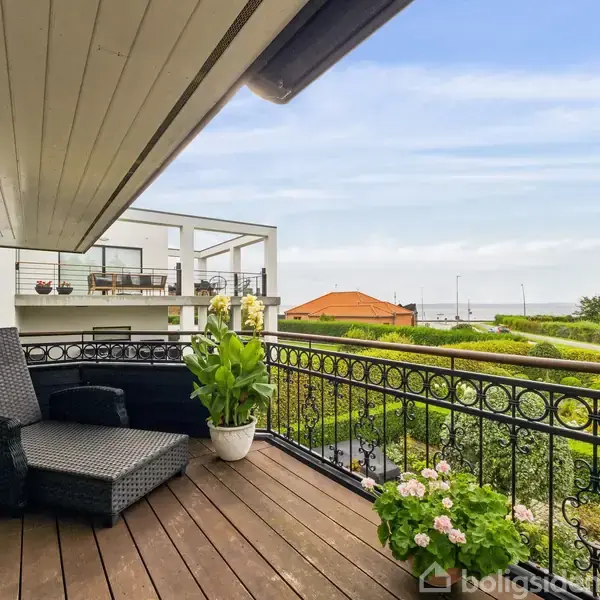 Terrassemøbler står på en træbelagt balkon, omkranset af smedejernsgelænder. Flere planter i potter langs kanten. I baggrunden ses en græsplæne og kystlinje under en klar himmel.