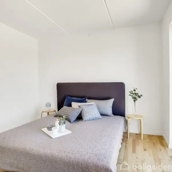 En seng med puder og et gråt tæppe står i et moderne, lyst soveværelse med trægulv. Ved siden af står natborde med en lampe og en plante.