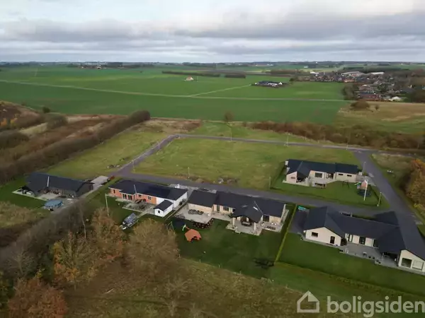 En række moderne huse ligger langs en vej. De ligger i et grønt landskab omgivet af marker og spredte træer under en overskyet himmel.