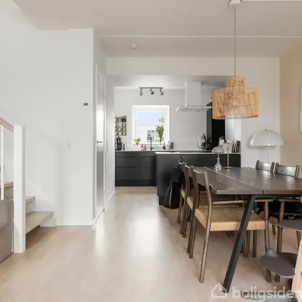Spisebord med stole står i et lyst, moderne køkken-alrum. Trappe til venstre, lyse trægulve, hvide vægge, og to billeder på endevæggen. Køkkenet har mørke skabe og er veludstyret.