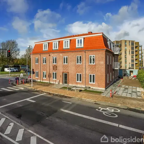 Rødt murstenshus står på hjørne af gade, omgivet af vejafmærkninger til cykelsti og fodgængerovergang, i et bymiljø med andre bygninger i baggrunden under blå himmel med skyer.