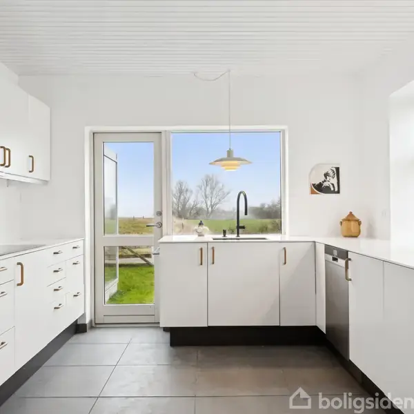 Et moderne køkken med hvide skabe og sorte håndtag. Rummet har en vask under et stort vindue med udsigt til en grøn have. Fliser dækker gulvet.