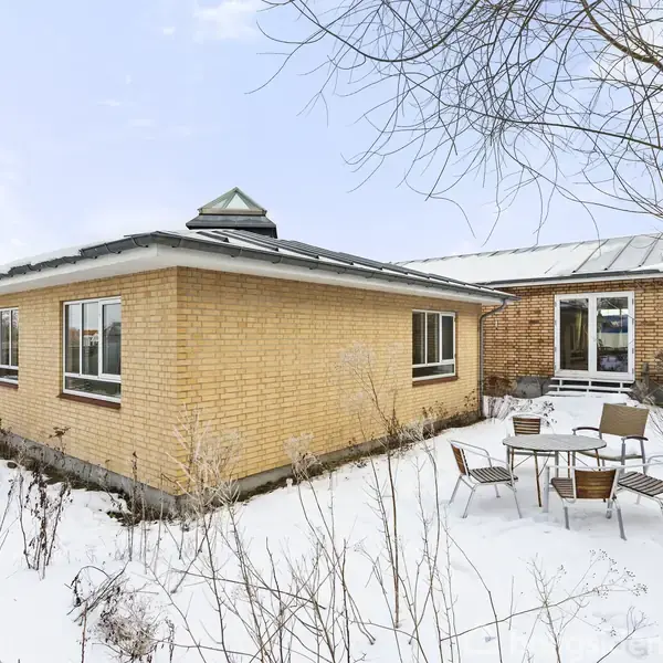 Hus i gule mursten står stille i en snedækket have. Ruderne er klare, og der er et lille bord med stole i haven. Træer uden blade omgiver stedet.