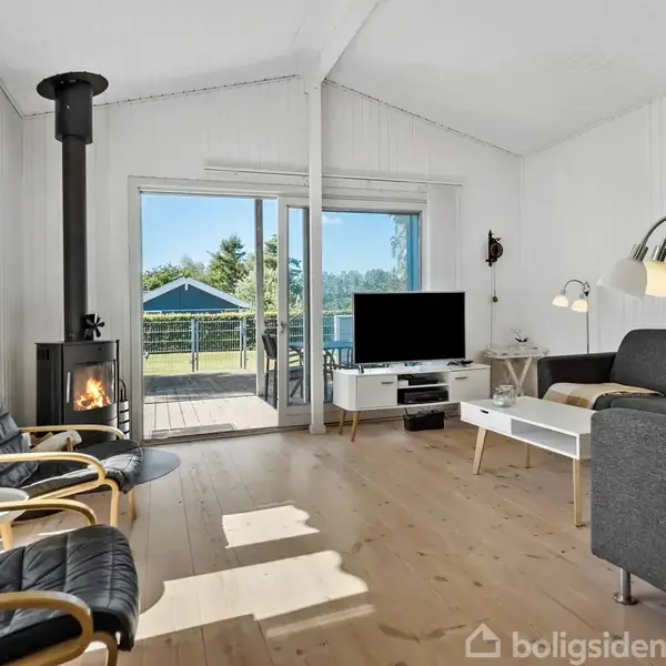 Brændeovn opvarmer hyggelig stue med lyse trægulve, sort sofa, fladskærms-tv, og to lænestole. Store glasdøre åbner til terrasse med udsigt til en have med træer.