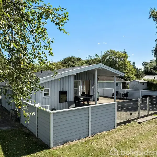 Et lyst træhus med stor terrasse står i en grøn have med træer omkring. Terrassemøbler er placeret under et halvtag. Himlen er klar og blå.