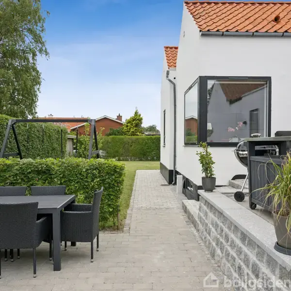 Et moderne hus med hvid facade og rødt tag står i en have. En terrasse har et udendørs spisebord med stole. Græsplæne og hække omgiver området.