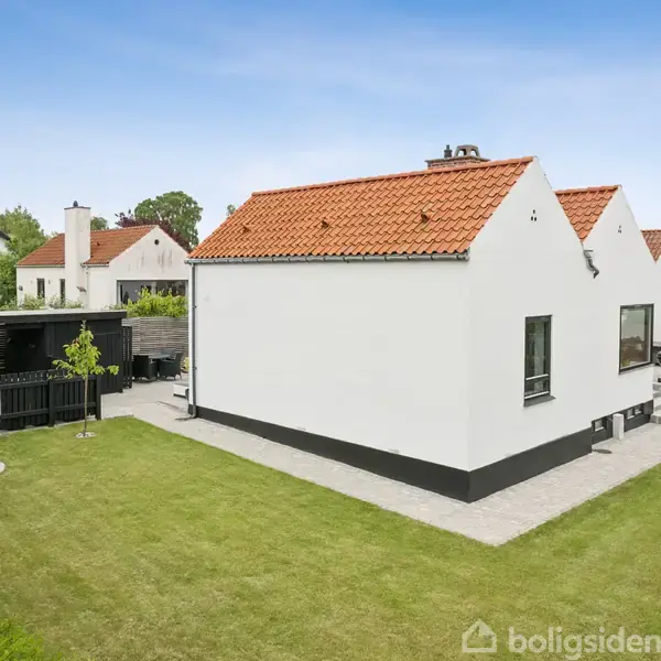 Et hvidt hus med rødt tegltag står i en velplejet have med en flagstang. Huset har sorte vinduer og en sort stenterrasse. Omkringliggende grønne buske ses.