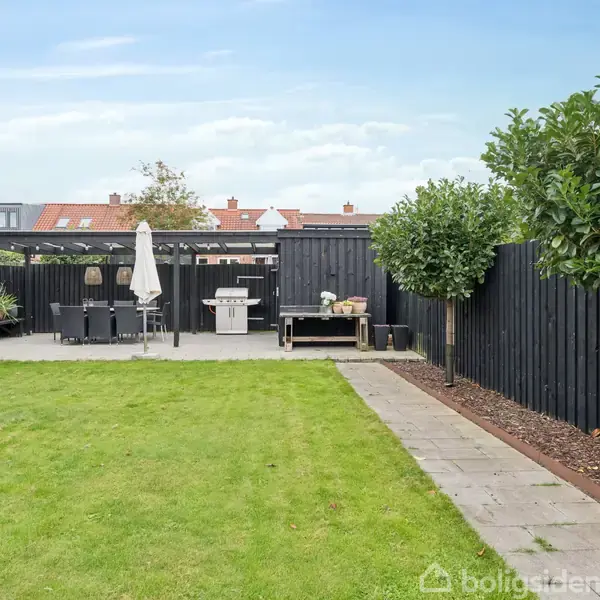 Græsplæne fører til en terrasse med sorte plankeværker og havemøbler under en pergola; omgivet af træer og buske i en villahave.