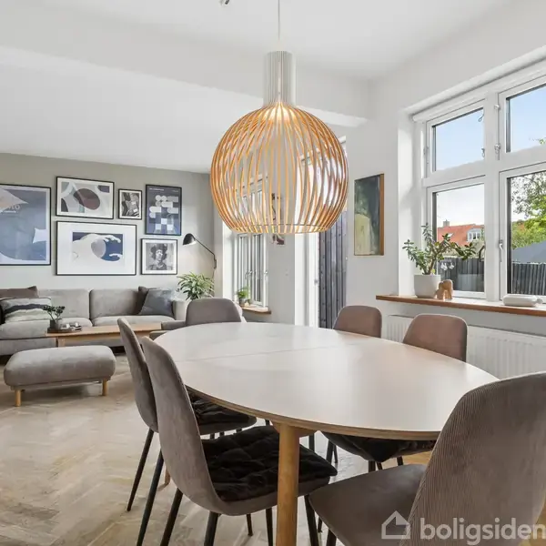 En oval spisebord med seks stole står under en dekorativ hængelampe. Ved siden af ses en grå sofa med puder og kunst på væggen. Stuen har lyst trægulv og store vinduer.