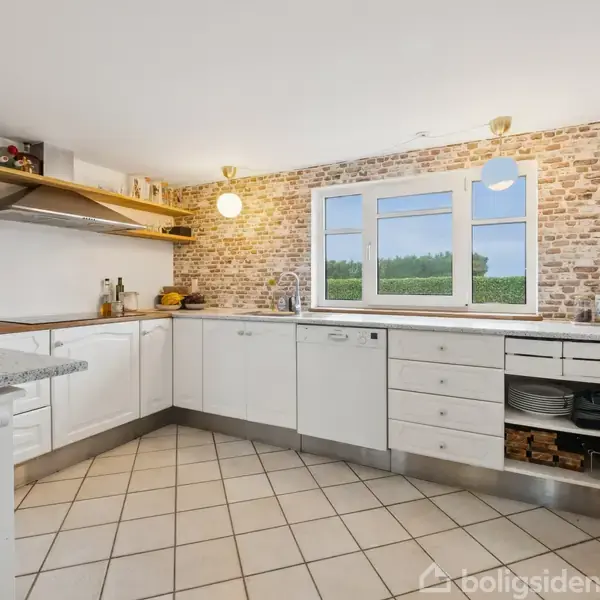 Et lyst køkken har hvide skabe, en opvaskemaskine og åbne hylder med glas og køkkenudstyr. En murstensvæg har et vindue med udsigt til grøn natur.