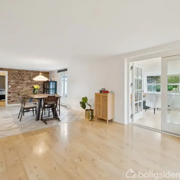 En stue med lyst trægulv indeholder et spisebord med stole. Til venstre ses en murstensvæg, og til højre åbner franske døre ud til endnu et rum med store vinduer.