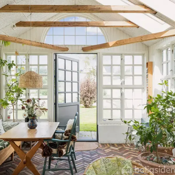 En hyggelig udestue med træmøbler og grønne planter. Sollys strømmer ind gennem mange vinduer. En åben dør fører ud til en frodig have.