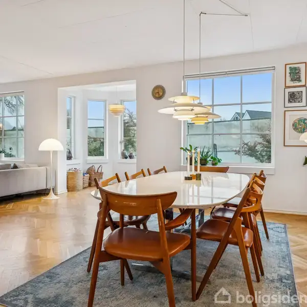 Ovalt spisebord med træstole står på et tæppe under en moderne lysekrone. I baggrunden, en stue med grå sofa, reol med bøger, og store vinduer, der fremhæver rummets lyse atmosfære.