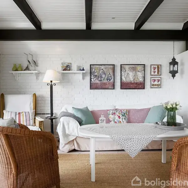 Stue med en hvid sofa fyldt med puder, foran et rundt bord med en dug. To kurvestole står foran, omgivet af en lampe, hylder og vægdekorationer i et lyst rum.