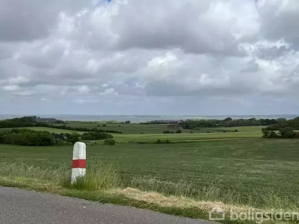 En vejpæl står ved vejkanten, omgivet af grønne marker under en overskyet himmel.