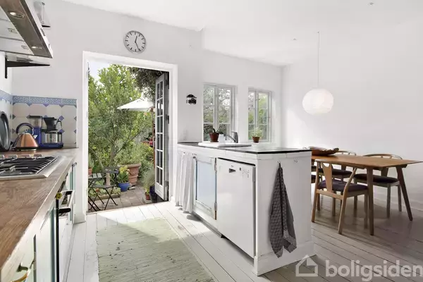 Køkken med hvidt interiør og træbord, vask i midten, åbne døre til terrasse med planter og parasol. Vægur hænger over døren, spisebord ved vindue.