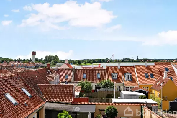 Røde tegltage dækker husene i en lille by. Sollys skinner på murene, mens træer og marker strækker sig i baggrunden under en blå himmel med enkelte skyer.