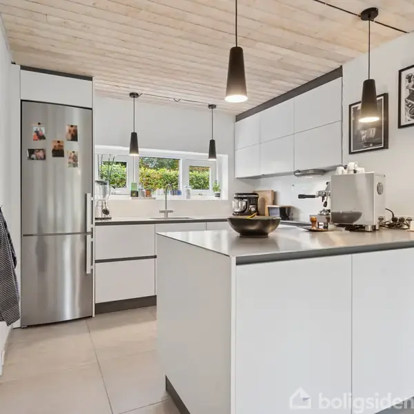 Et moderne køkken har hvide skabe, en stål-fryser og køleskab. Der er en kaffemaskine og nogle bøger på køkkenbordet. Vinduer i baggrunden giver udsigt til grønne omgivelser.