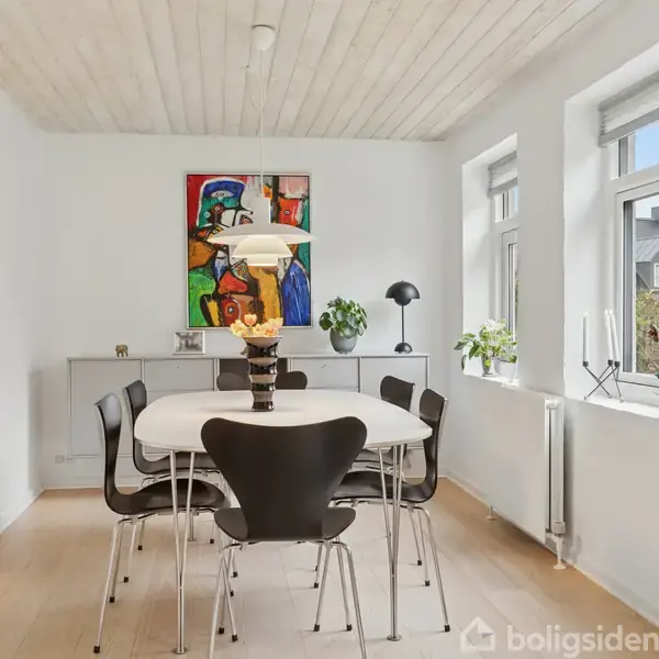 Et spisebord med seks sorte stole omkring. Bordet har en moderne lampe. Værelset har hvidmalede vægge, vinduer til højre, planter og kunst på væggene.